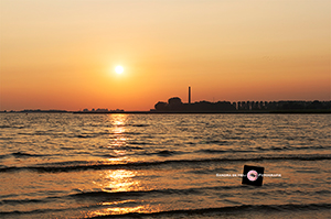 Oranje zonsondergang bij een rustig kabbelend IJsselmeer. Op de achtergrond is de schoorsteen van het Ir. Woudagemaal te zien.
